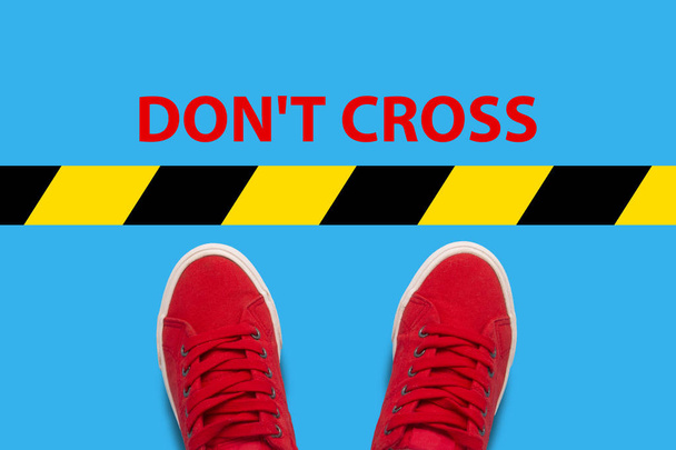 Vrouwelijke voeten in rode sneakers staande voor een waarschuwing zwart-en-gele lijn met tekst oversteken niet. Concept van de vernietiging van grenzen en schendingen van de regels. Plat lag, top uitzicht. - Foto, afbeelding