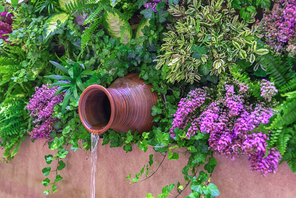 Vertical gardening in landscape design. Wall-mounted jug - a waterfall in the thickets of green plants and lilac branches - Photo, Image