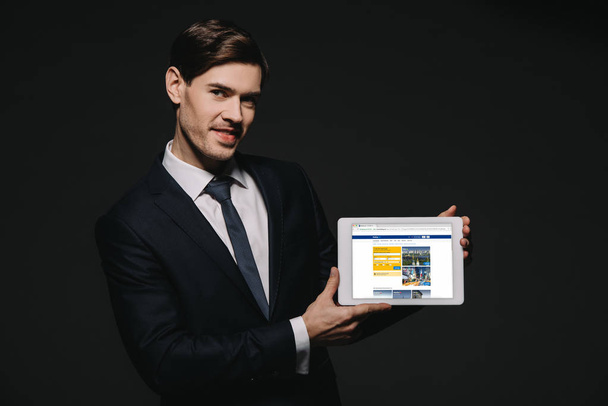 cheerful businessman holding digital tablet with booking.com app in screen isolated on black - Foto, immagini