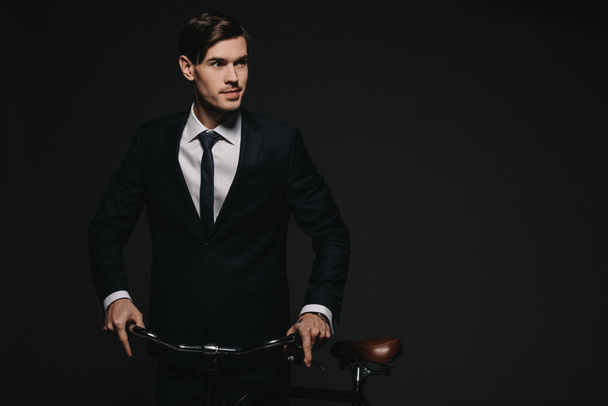 handsome businessman in suit holding bicycle isolated on black - Foto, afbeelding