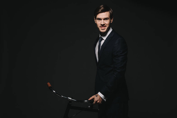 smiling businessman in suit holding bicycle isolated on black - Φωτογραφία, εικόνα