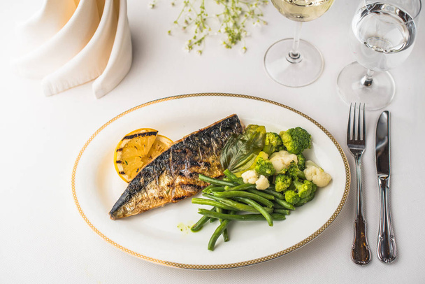Grilled fish and steamed vegetables served on plate on white table in restaurant - Фото, изображение