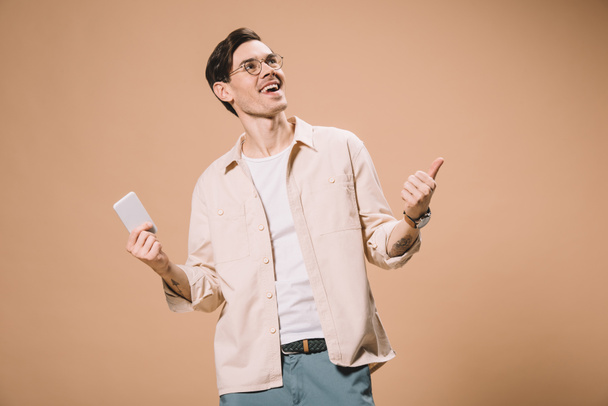 hombre alegre en gafas sosteniendo teléfono inteligente y mostrando el pulgar hacia arriba aislado en beige
  - Foto, imagen