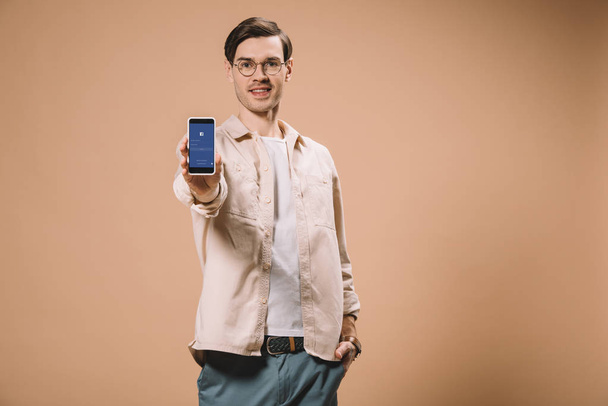 cheerful man standing with hand in pocket and holding smartphone with facebook app on screen isolated on beige  - Photo, Image