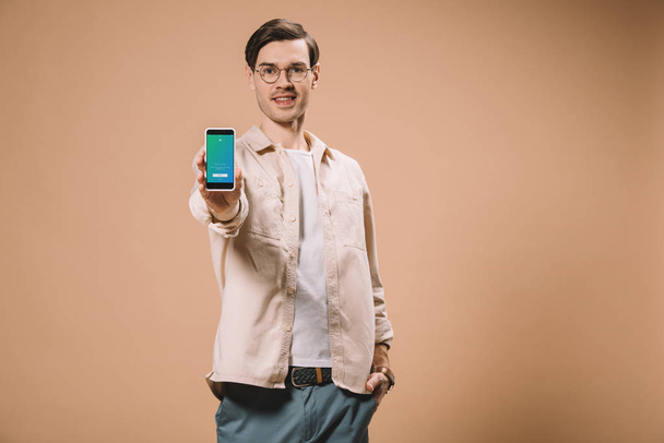 cheerful man standing with hand in pocket and holding smartphone with twitter app on screen isolated on beige  - Fotografie, Obrázek