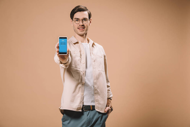 веселый человек, стоящий с рукой в кармане и держа смартфон с приложением Skype на экране изолированы на бежевый
  - Фото, изображение