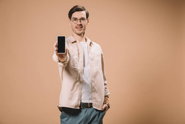 handsome man standing with hand in pocket and holding smartphone with blank screen isolated on beige  - 写真・画像