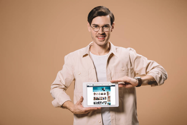 handsome man in glasses holding digital tablet with amazon app on screen isolated on beige  - Zdjęcie, obraz