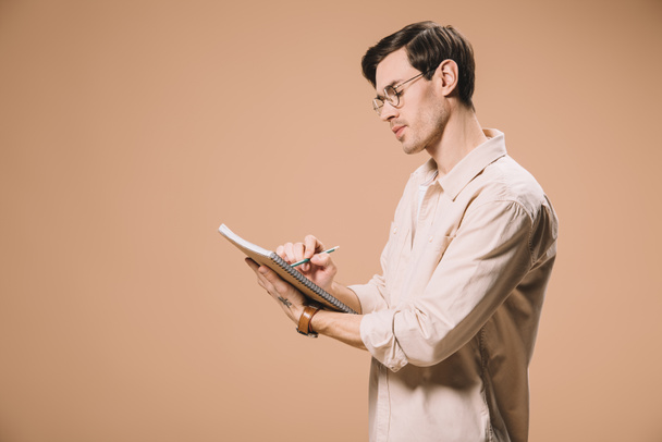 thoughtful man in glasses writing notebook isolated on beige  - Foto, Imagem