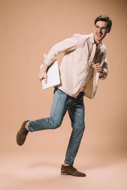 happy man in glasses running with laptop on beige background  - Фото, изображение