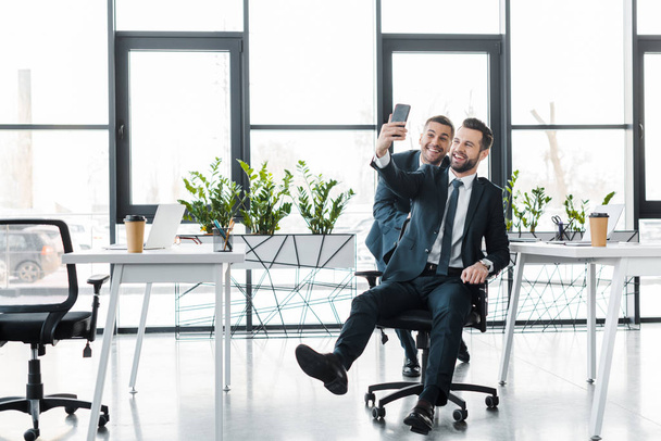busimessmen guapos tomando selfie y sonriendo en oficina moderna
  - Foto, Imagen