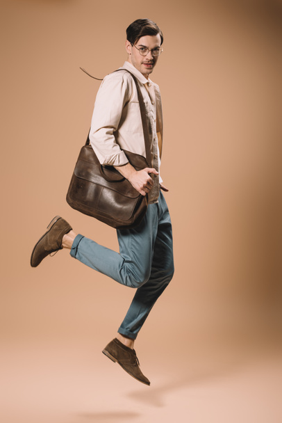 handsome man in glasses jumping with bag on beige background  - Zdjęcie, obraz