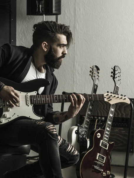 Photo of a man with beard sitting and playing his electric guitar in a recording studio. - Zdjęcie, obraz