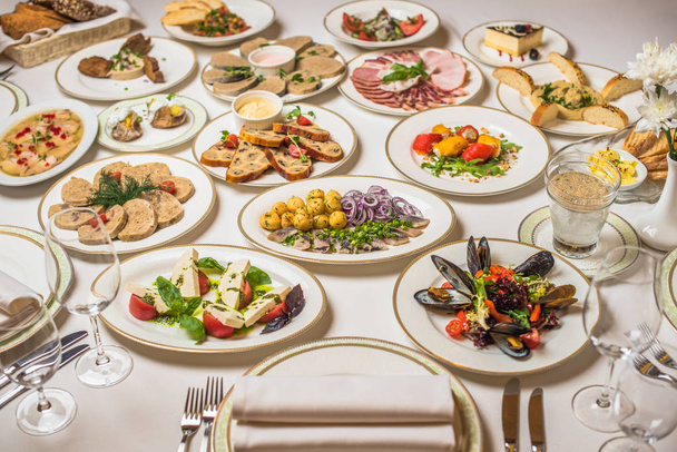 Restaurace tabulka s mnoha různých pokrmů. Talíře, vidličky, nože, sklenice na bílý ubrus. Květiny ve váze - Fotografie, Obrázek