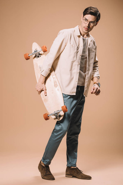 man in glasses holding wooden skateboard while standing on beige background - Photo, Image