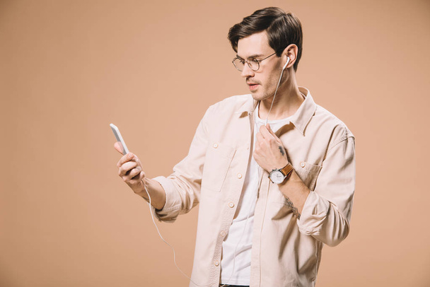 красивий чоловік в окулярах дивиться на смартфон під час прослуховування музики в навушниках ізольовано на бежевому
  - Фото, зображення