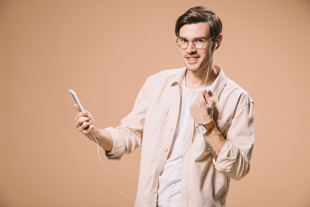 smiling man in glasses holding smartphone and listening music in earphones isolated on beige  - Foto, imagen