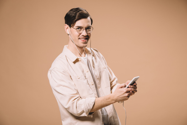 fröhlicher Mann mit Brille, Smartphone in der Hand und Musik im Kopfhörer isoliert auf Beige hörend  - Foto, Bild