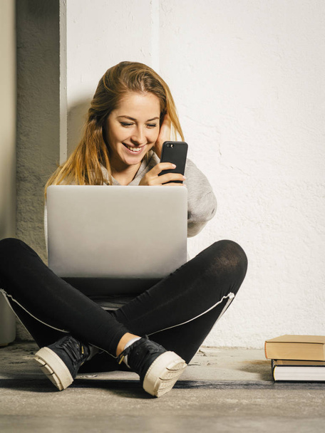 Foto van een jonge vrouw in een hal met een laptop zitten en lachen om een bericht op haar smartphone. - Foto, afbeelding