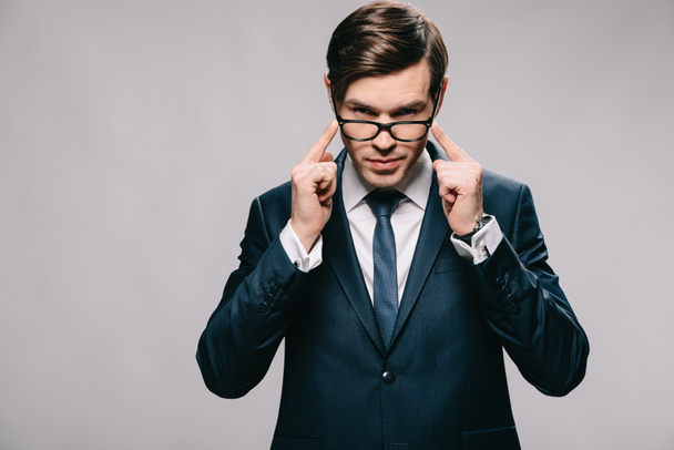 confident businessman holding glasses while standing in suit isolated on grey - Фото, изображение
