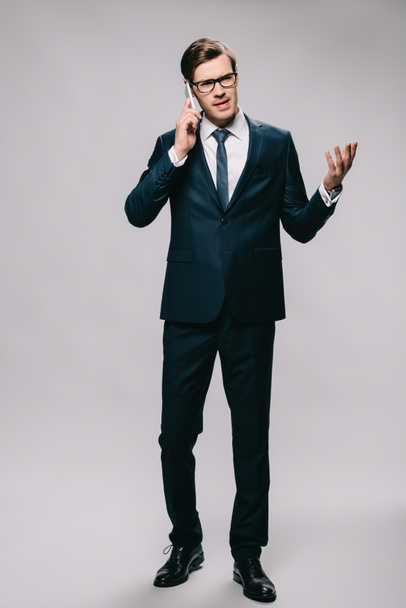 handsome businessman talking on smartphone while standing in suit on grey background  - Fotó, kép