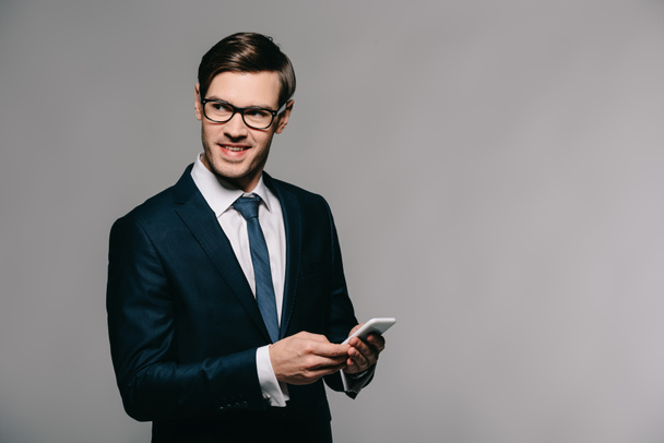 веселый бизнесмен, использующий смартфон, стоя в костюме на сером фоне
  - Фото, изображение