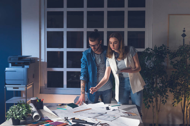 Dois jovens arquitetos caucasianos, um homem e uma mulher, usando um tablet enquanto trabalham juntos em um escritório à noite
. - Foto, Imagem