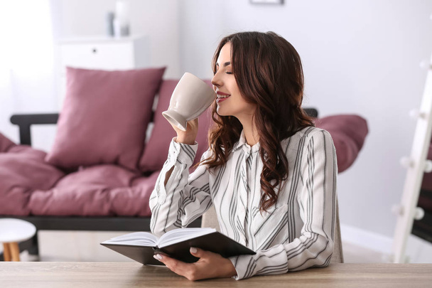 Beautiful young woman drinking coffee while reading book at home - Photo, Image