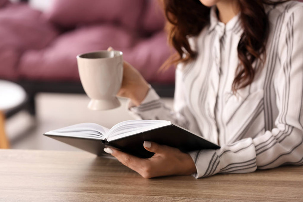 Όμορφη νεαρή γυναίκα που πίνει καφέ, διαβάζοντας το βιβλίο στο σπίτι - Φωτογραφία, εικόνα