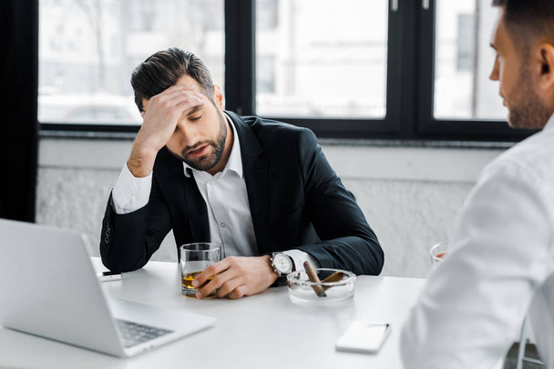 selective focus of tired businessman having headache while holding glass with alcohol drink in modern office  - Photo, Image