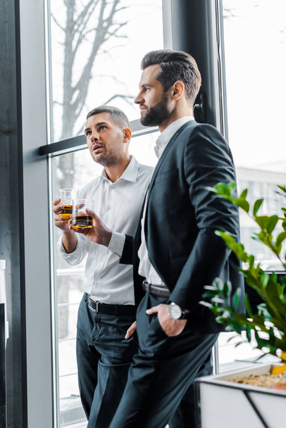 bearded businessman standing with hand in pocket and holding glass of whiskey near coworker - Photo, Image