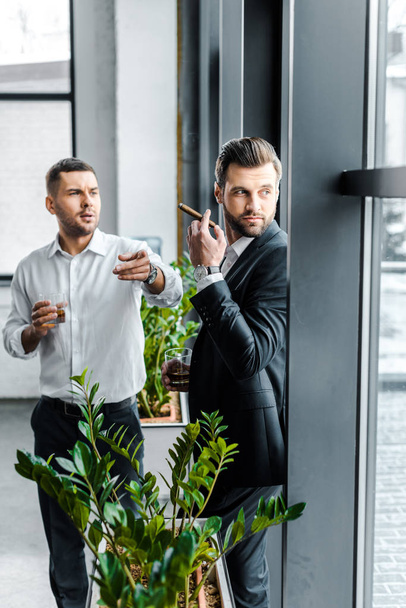 businessman pointing with finger while holding glass of whiskey near coworker with cuban cigar - Photo, Image