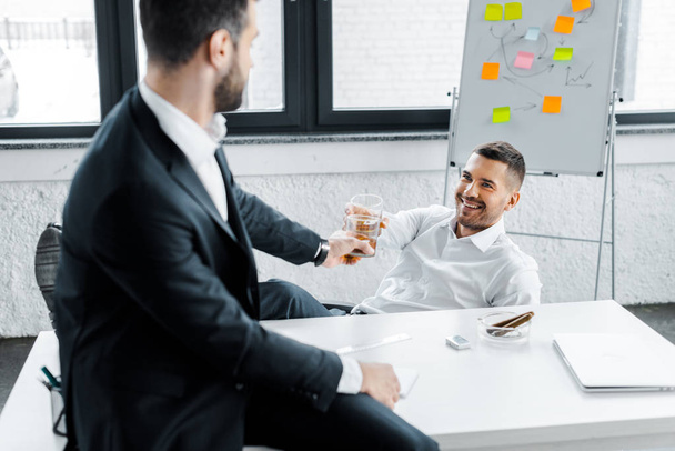 cheerful businessman clinking with alcohol drink in glass with coworker in modern office  - Fotó, kép