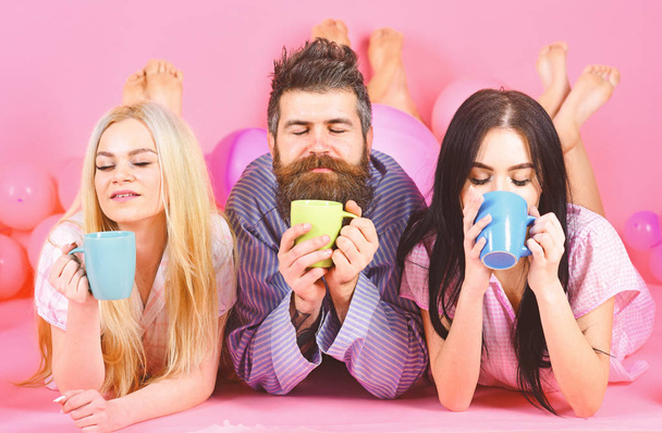 Lovers in love drink coffee in bed. Threesome relax in morning with coffee. Lovers concept. Man and women, friends on sleepy faces lay, pink background. Man and women in domestic clothes, pajamas - Foto, Imagem