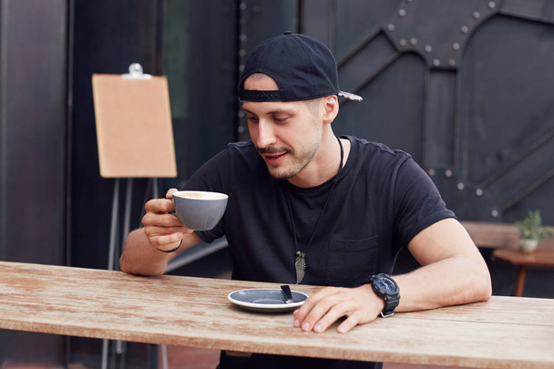 Menschen und Lebensstil-Konzept. attraktive erfolgreiche junge Hipster Kaffee trinken sitzt am Café-Tisch mit Becher, blickt auf Tasse mit foxy Lächeln, in der Hoffnung, dass harte Getränke wecken ihn auf. Hintergrund-Attrappe. - Foto, Bild