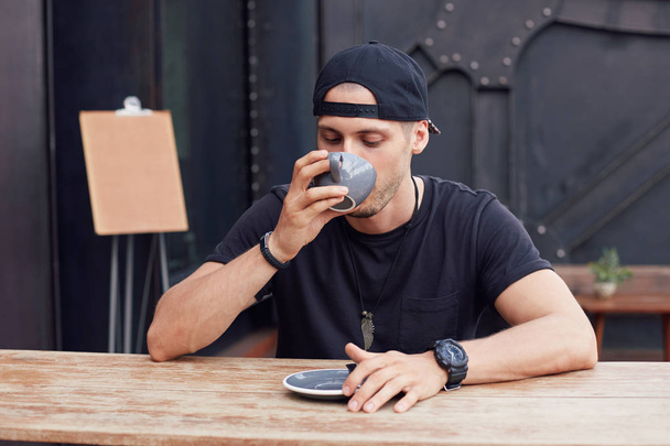 Sleepy Caucasian stylish student holding mug with fresh cappuccino, drinking fast not to be late at university, sitting outdoors, dressed in summer clothes, cafeteria menu mock up on background.  - Foto, imagen