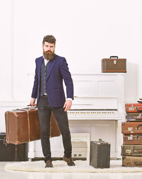 Man, traveller with beard and mustache with luggage, luxury white interior background. Luggage and vacation concept. Macho elegant on strict face stands near pile of vintage suitcase, holds suitcase - Foto, immagini