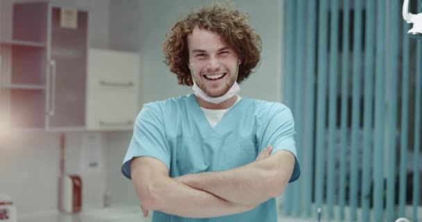 Portrait of a smiling young man surgeons or doctor putting on surgical mask and looking straight to the camera background modern clinic room. shot on red epic - Metraje, vídeo