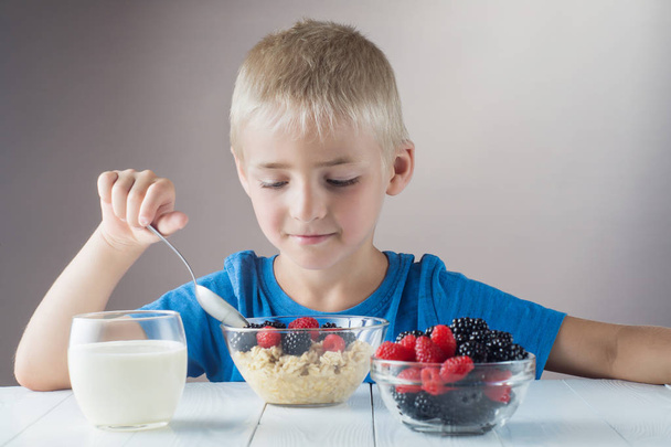 Little boy is eating oatmeal with berries. The concept of a healthy breakfast for children, porridge with raspberries and blackberries, a glass of yogurt on white wooden table. - Photo, Image