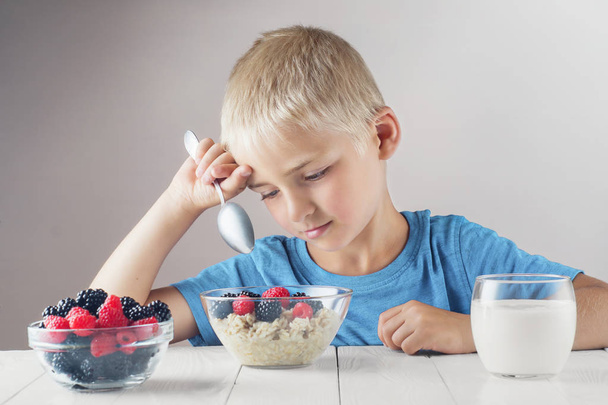 Sad child does not want to eat porridge with berries. Little boy eats without appetite.  The concept of a healthy breakfast for children, porridge with raspberries and blackberries, a glass of yogurt. - Φωτογραφία, εικόνα