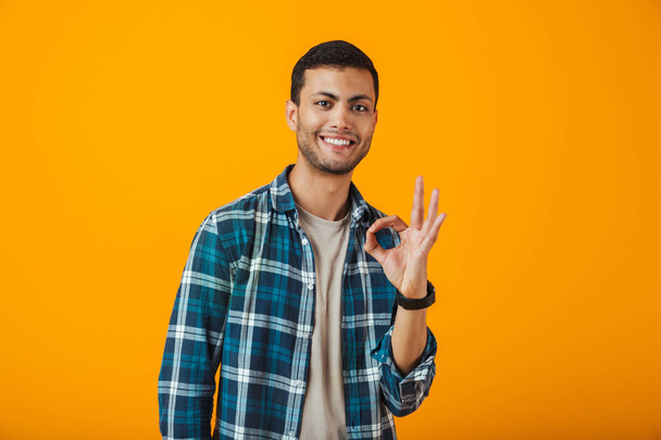 Cheerful young man wearing plaid shirt standing isolated over orange background, showing ok - Foto, Bild