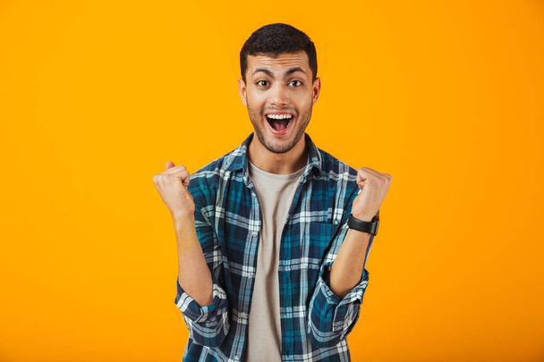 Cheerful young man wearing plaid shirt standing isolated over orange background, celebrating - 写真・画像