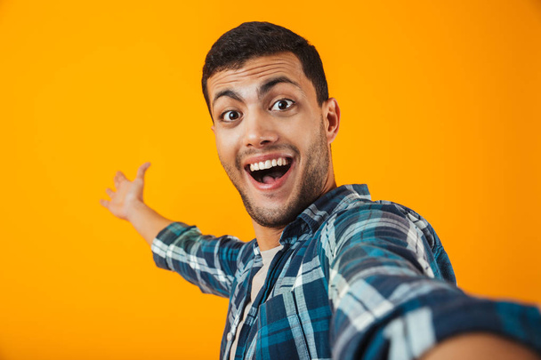 Cheerful young man wearing plaid shirt standing isolated over orange background, taking a selfie - Foto, Bild