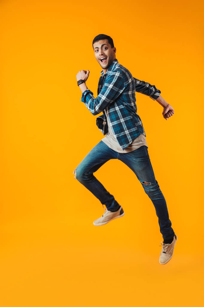 Full length portrait of a happy young man wearing plaid shirt isolated over orange background, jumping - Foto, imagen