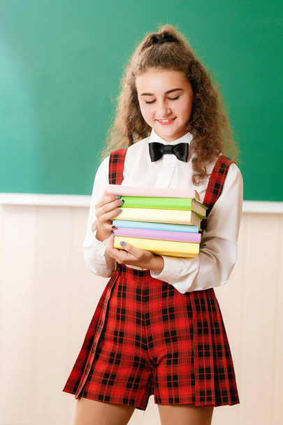 beautiful schoolgirl stands in the classroom and holds books against the background of the blackboard. - Photo, image
