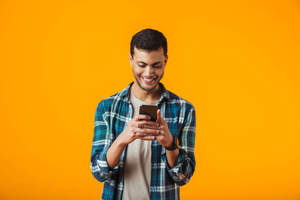 Cheerful young man wearing plaid shirt standing isolated over orange background, using mobile phone - 写真・画像