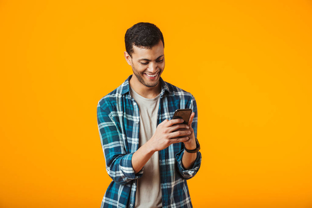 Cheerful young man wearing plaid shirt standing isolated over orange background, using mobile phone - Фото, зображення