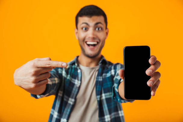 Cheerful young man wearing plaid shirt standing isolated over orange background, showing blank screen mobile phone - Fotoğraf, Görsel