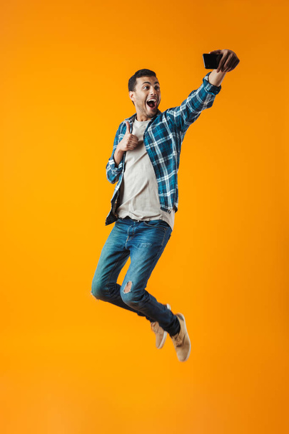 Full length portrait of a happy young man wearing plaid shirt isolated over orange background, jumping, taking a selfie - Foto, Imagem