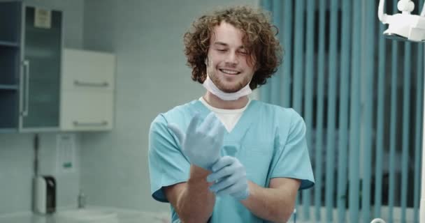 Young charismatic dentist or surgeons in front of the camera very happy and smiling in a hospital room , putting his surgical blue gloves and getting ready to start the operation. 4k - Video